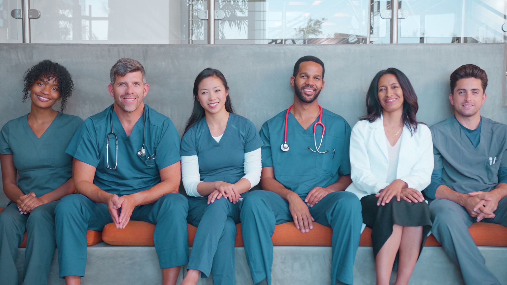 Portrait Of Multi-Cultural Medical Or Surgical Team Wearing Scrubs Sitting Inside Hospital Together